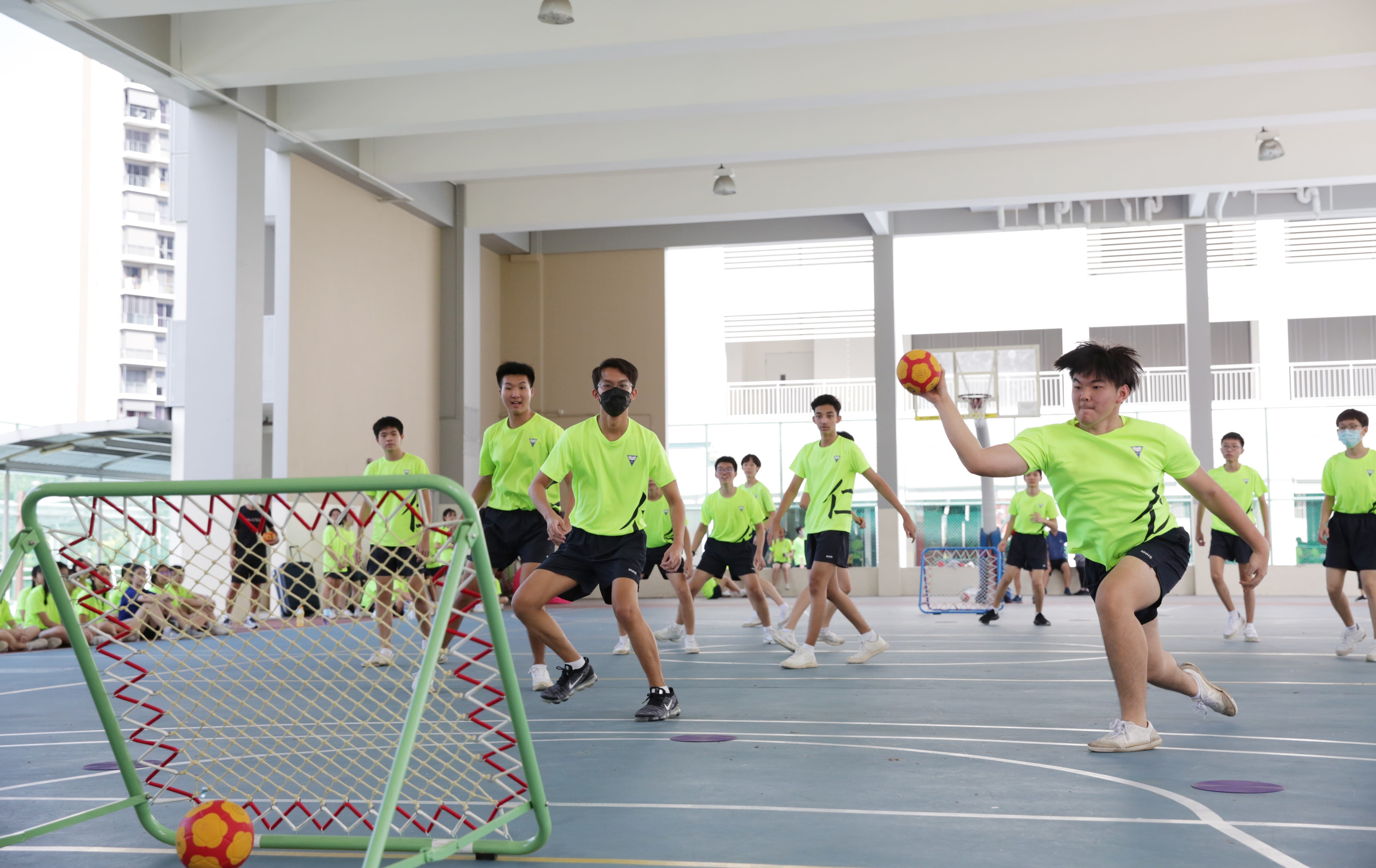 Students in neon green uniforms play handball on an indoor court, with one student throwing a red and yellow ball toward a small goal.
